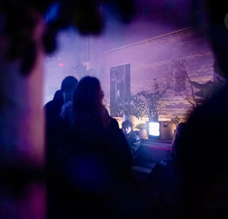 MARIAM KVASHILAVA EXHIBITIONeople sitting and standing in a dimly lit room with neon lights, plants, and a face on a small screen.