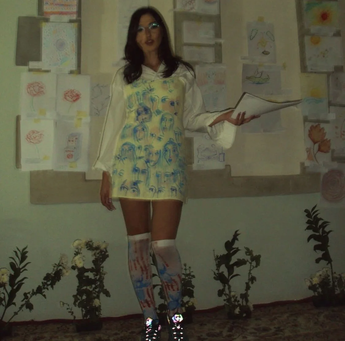 Person with long dark hair, glasses, wearing a cream-colored dress with face sketches, white thigh-high stockings with red and blue designs, and colorful shoes, standing in front of a wall with children's drawings. MARIAM KVASHILAVA EXHIBITION