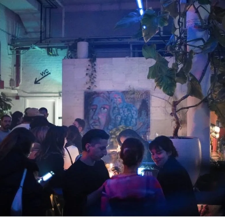People gathered in a dimly lit indoor space with blue and purple lighting, standing and talking near a large potted plant, with a mural of a woman on the wall in the background. A sign with an arrow points to the left labeled 'W.C.'. MARIAM KVASHILAV