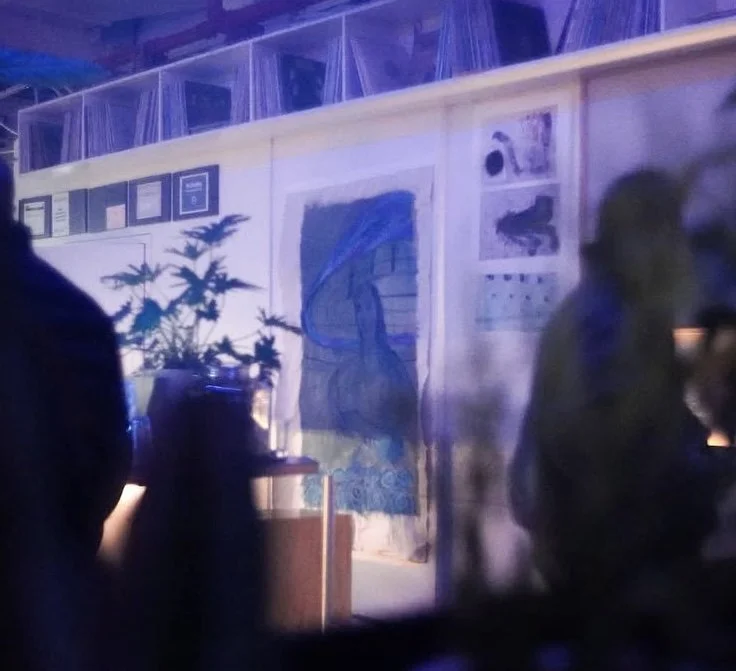 A blurry photo of an indoor space with artwork on the wall, a potted plant, and people in the foreground.