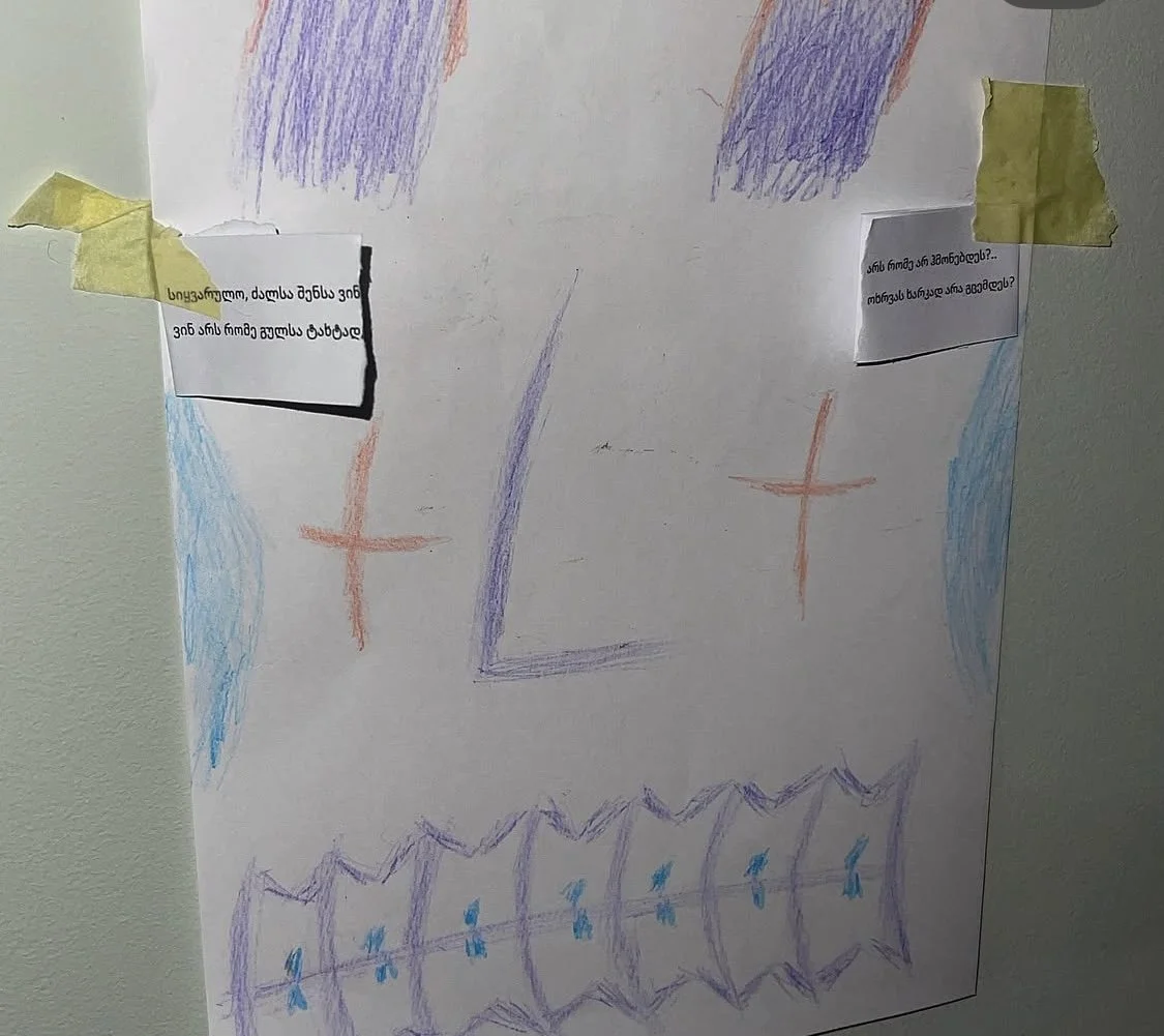 Children's drawing on white paper with colorful crayon scribbles, crosses, and patterns, taped to a wall with yellow tape. Two small paper notes with text are attached, one on each side.