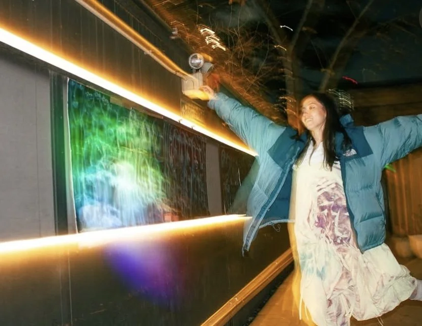 A woman with arms outstretched in a blue jacket and patterned dress, standing next to a colorful reflecting window at night. MARIAM KVASHILAVA EXHIBITION
