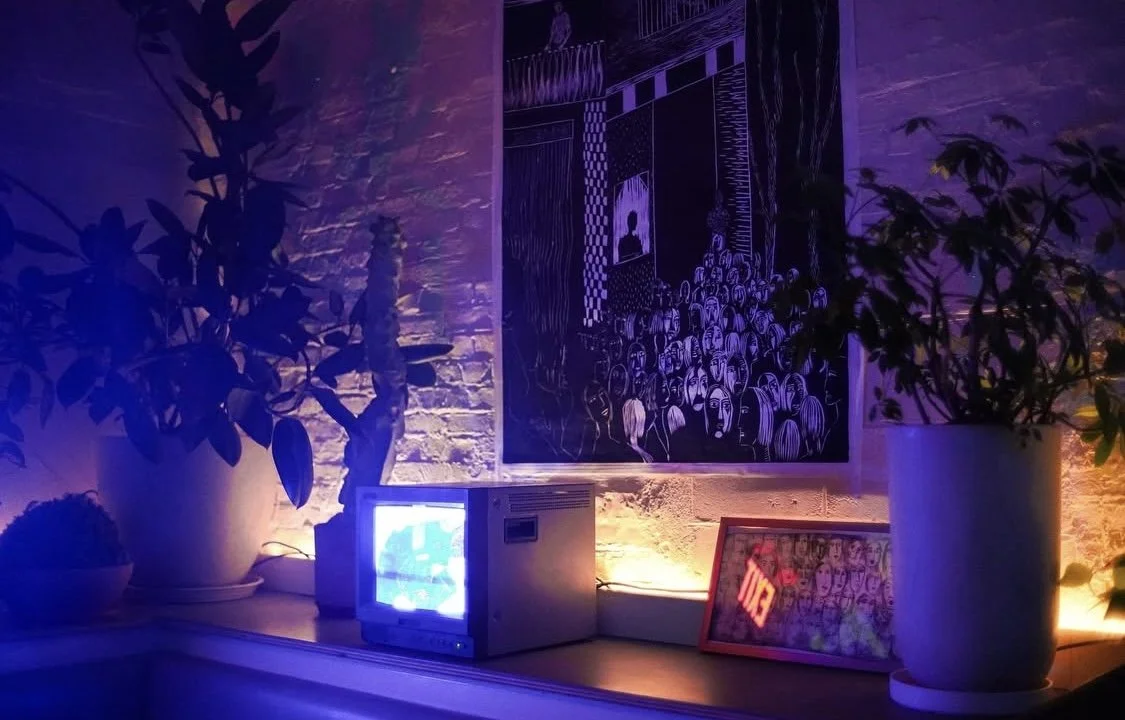 A small television, framed artwork, and potted plants are placed on a windowsill with purple and warm lighting, against a textured brick wall with a large black and white art piece depicting a crowd of people.