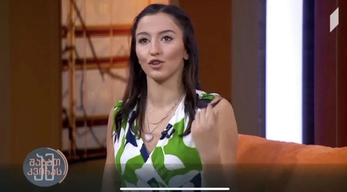 MARIAM KVASHILAVA with dark hair wearing a white, green, and black patterned sleeveless dress sitting on an orange couch during a TV interview.