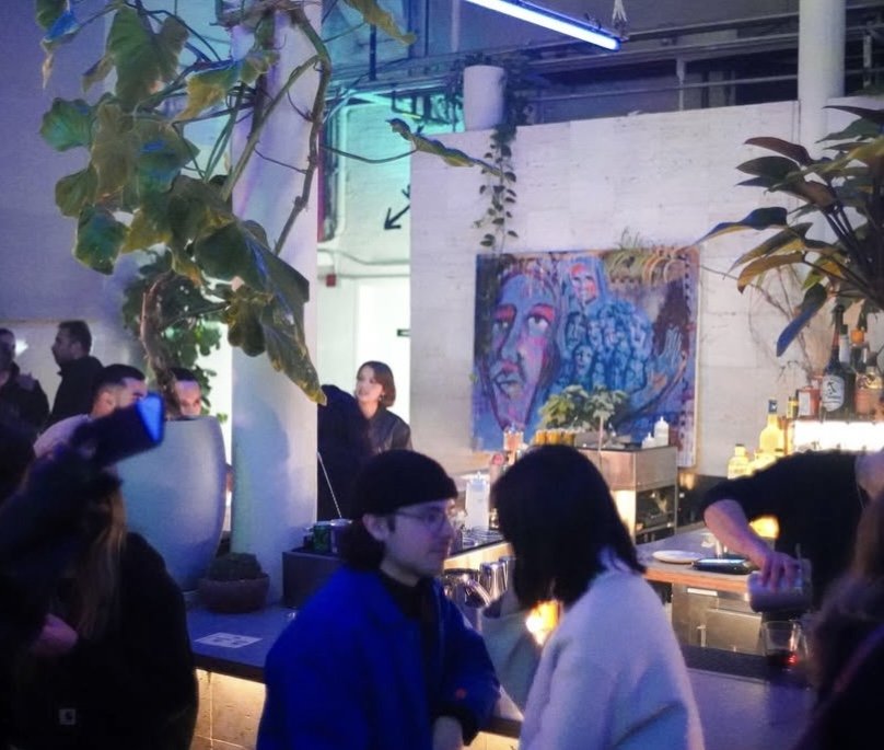 People socializing at a bar with a colorful abstract face painting on the wall, large indoor plants, and neon lighting.
