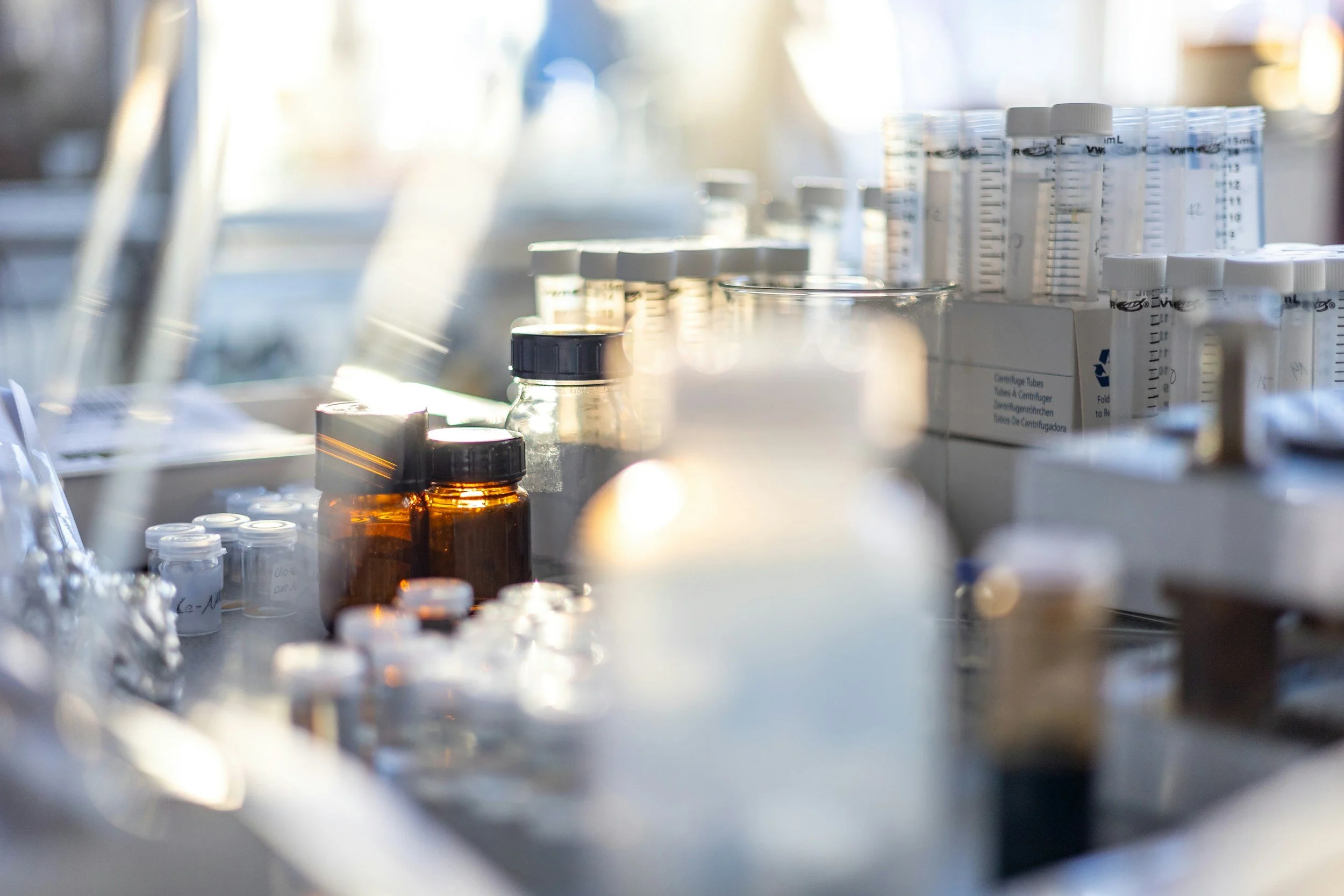 Close-up of a laboratory work station with various glass bottles and plastic containers, some with white labels and lids, on a workbench bathed in natural light.