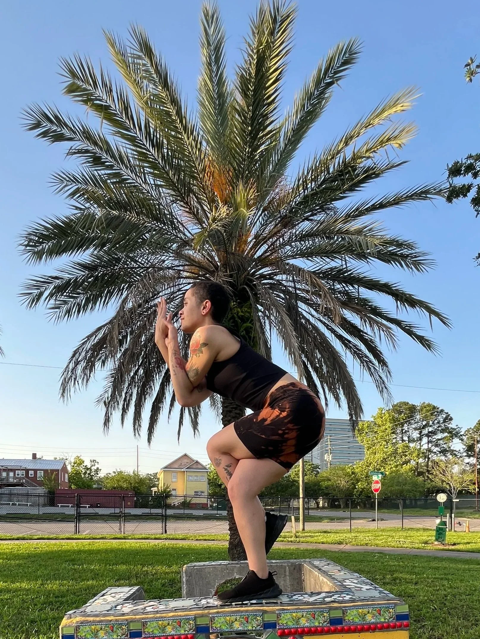A young person with a short afro haircut is standing in eagle pose in front of a palm tree in a park