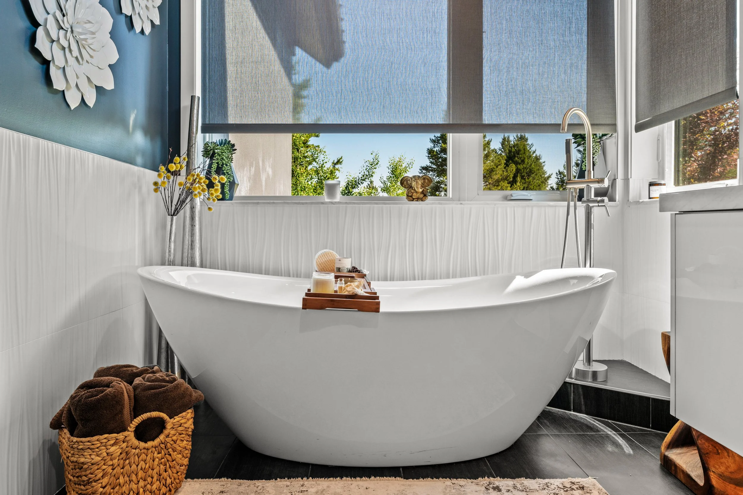 Modern bathroom with a white freestanding bathtub, a basket of brown towels, a tray with bath products, and large windows with views of green trees outside.