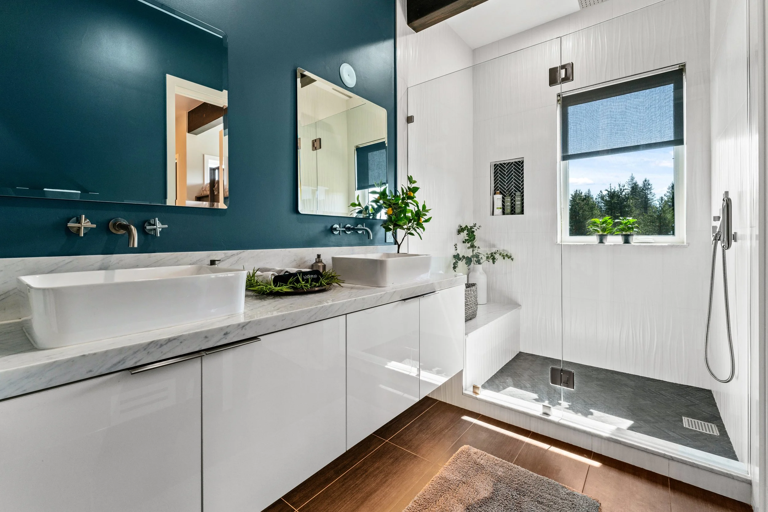 Modern bathroom with double white vessel sinks on a marble countertop, dark teal accent wall with two mirrors, white cabinetry, a walk-in shower with a glass door, window view of trees, and green plants