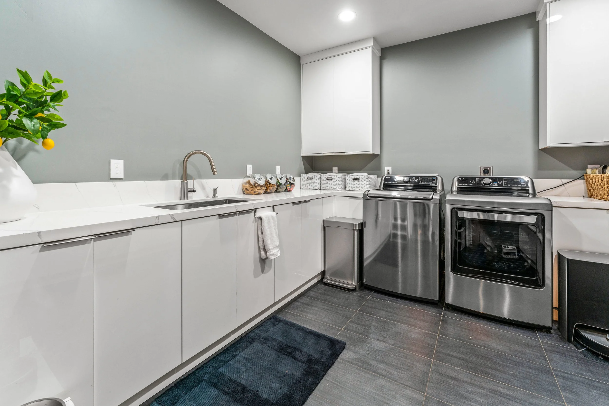 Modern laundry room with gray walls, white cabinets, dark tile floors, stainless steel washer and dryer, sink, and organized storage baskets.