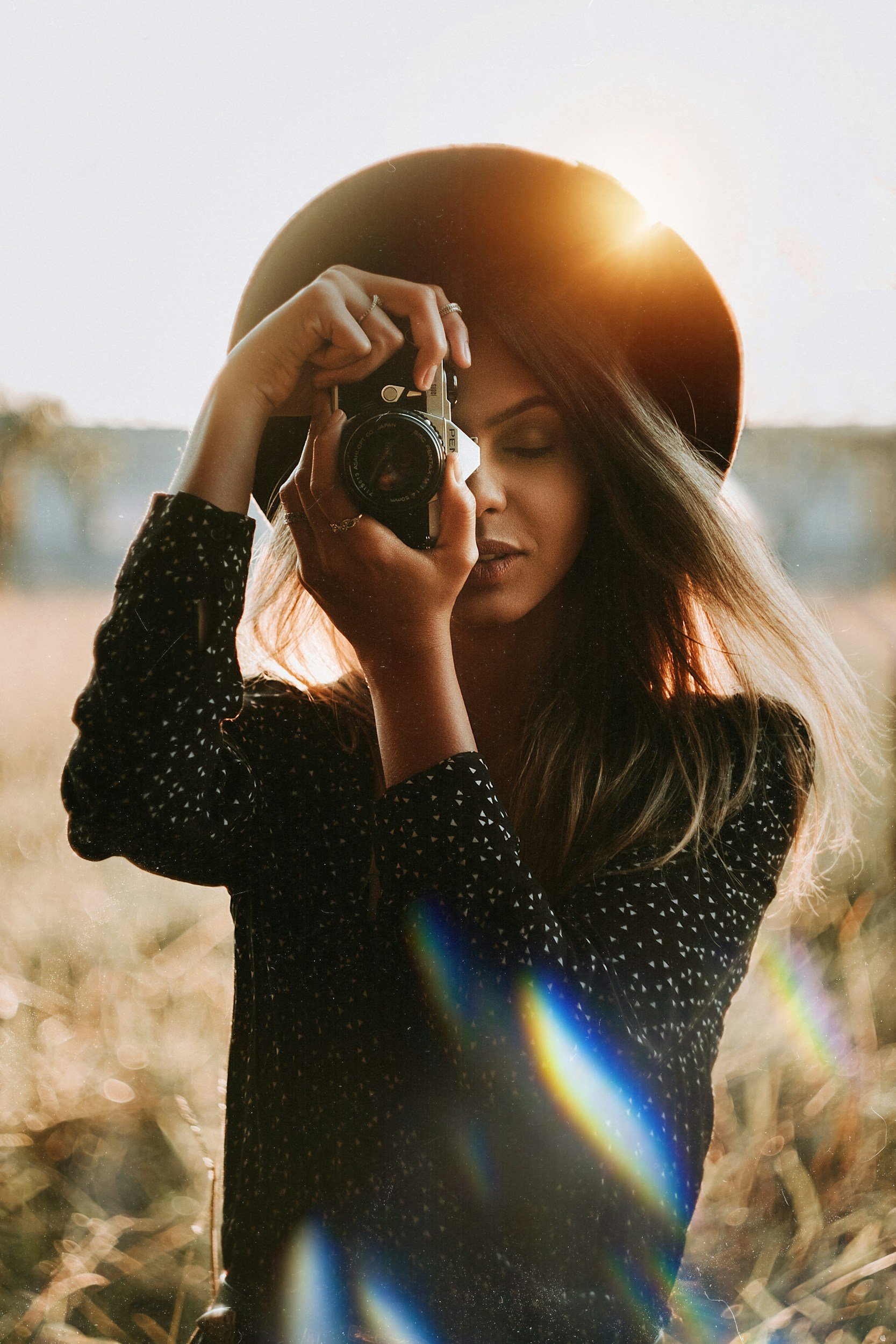 A woman with long hair wearing a black hat and a black polka-dot shirt is taking a photo with a camera outdoors at sunset, with sunlight and slight rainbow lens flare.