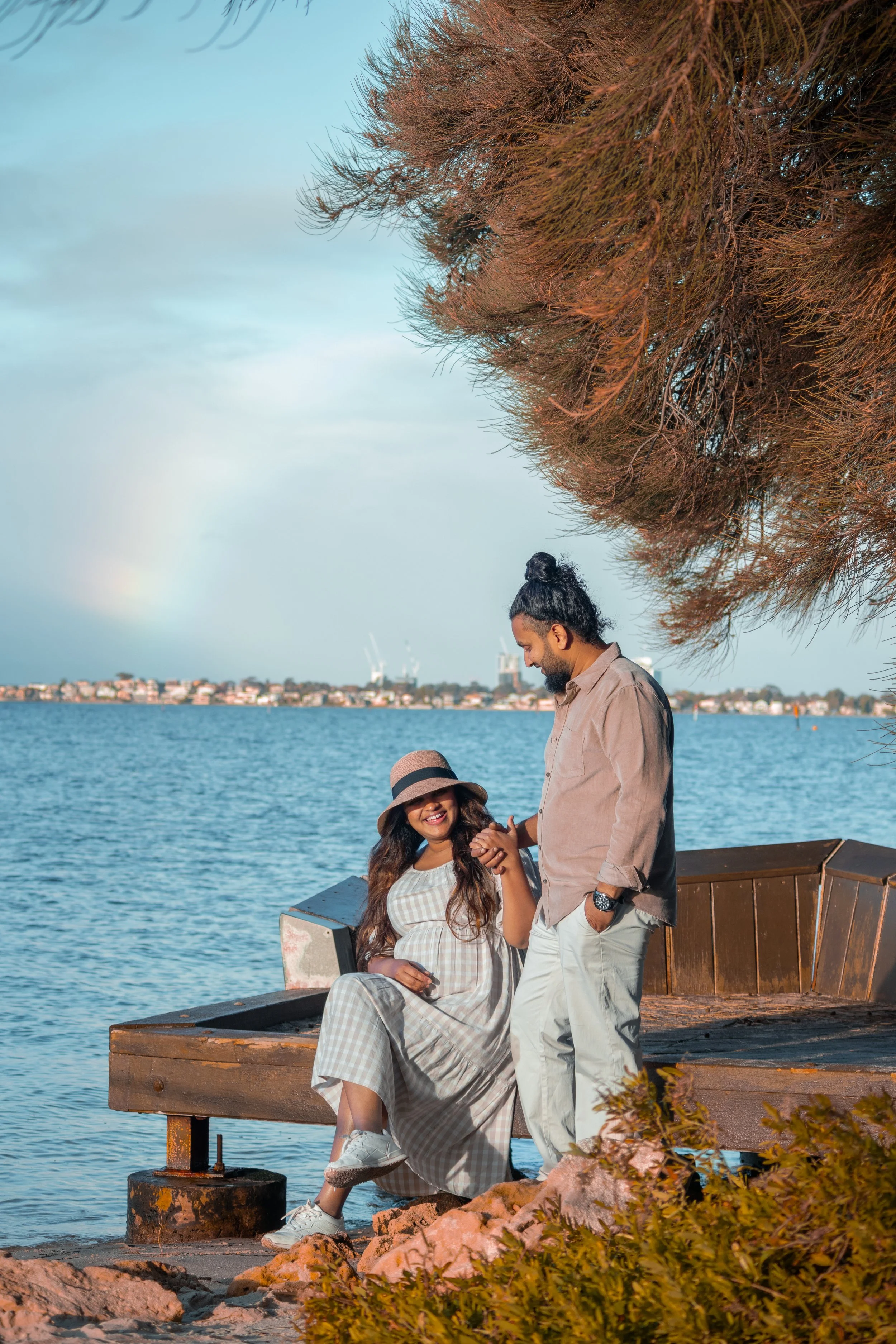 A couple by a lakeside with a woman sitting on a wooden bench and a man standing beside her, holding her hand, both smiling, with trees overhead and a distant cityscape with cranes and a rainbow in the sky.