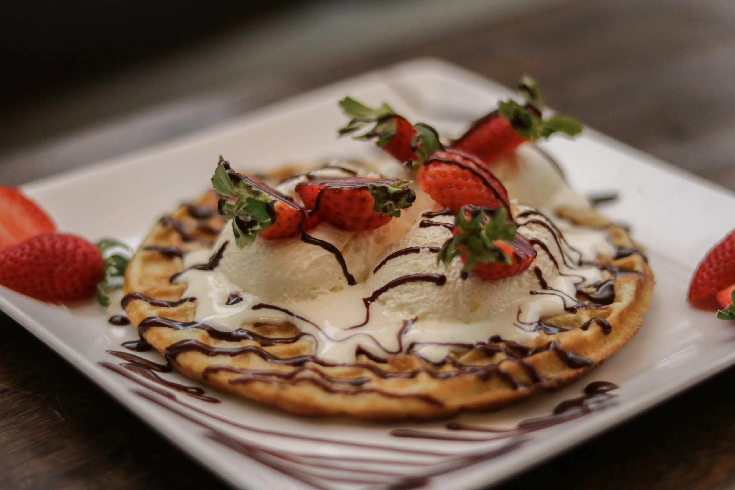 Waffle topped with vanilla ice cream, sliced strawberries, and chocolate drizzle, served on a white plate.