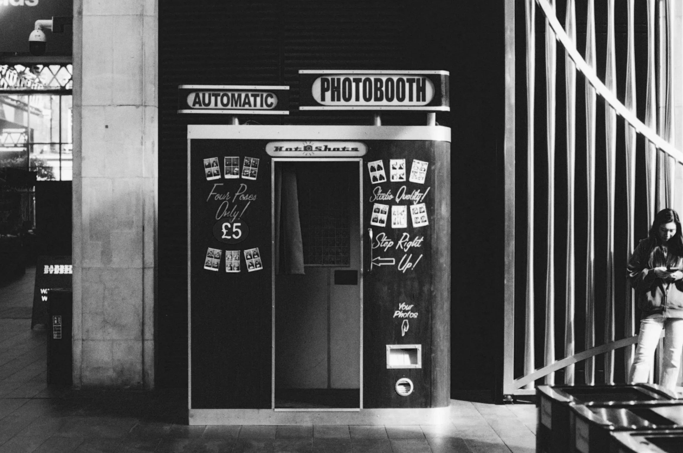 Photobooth kiosk with signs indicating 'Automatic,' 'Photobooth,' and pricing of £5 for four poses. The kiosk has a curtain entrance, and there are photo samples and instructions on the front. A woman is looking at her phone to the right of the kiosk, standing next to a metallic wall with vertical bars.