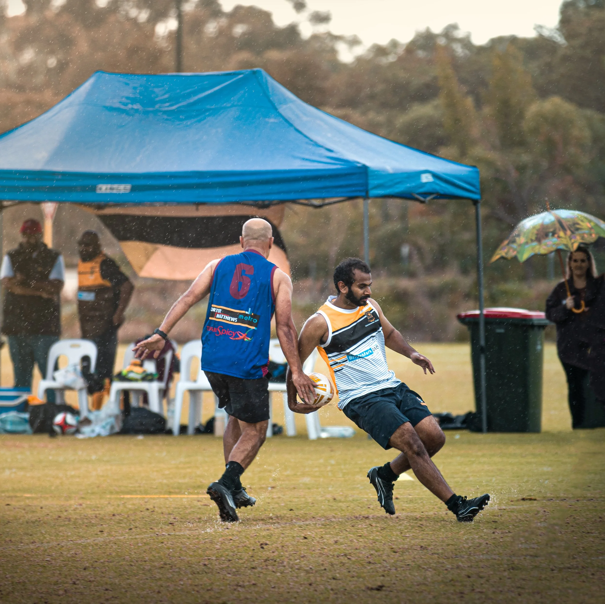 Two men playing rugby on a grassy field during rain. One is in a blue jersey with the number 6, and the other in a white, black, and gold jersey holding a rugby ball. A blue canopy and people with umbrellas are in the background.