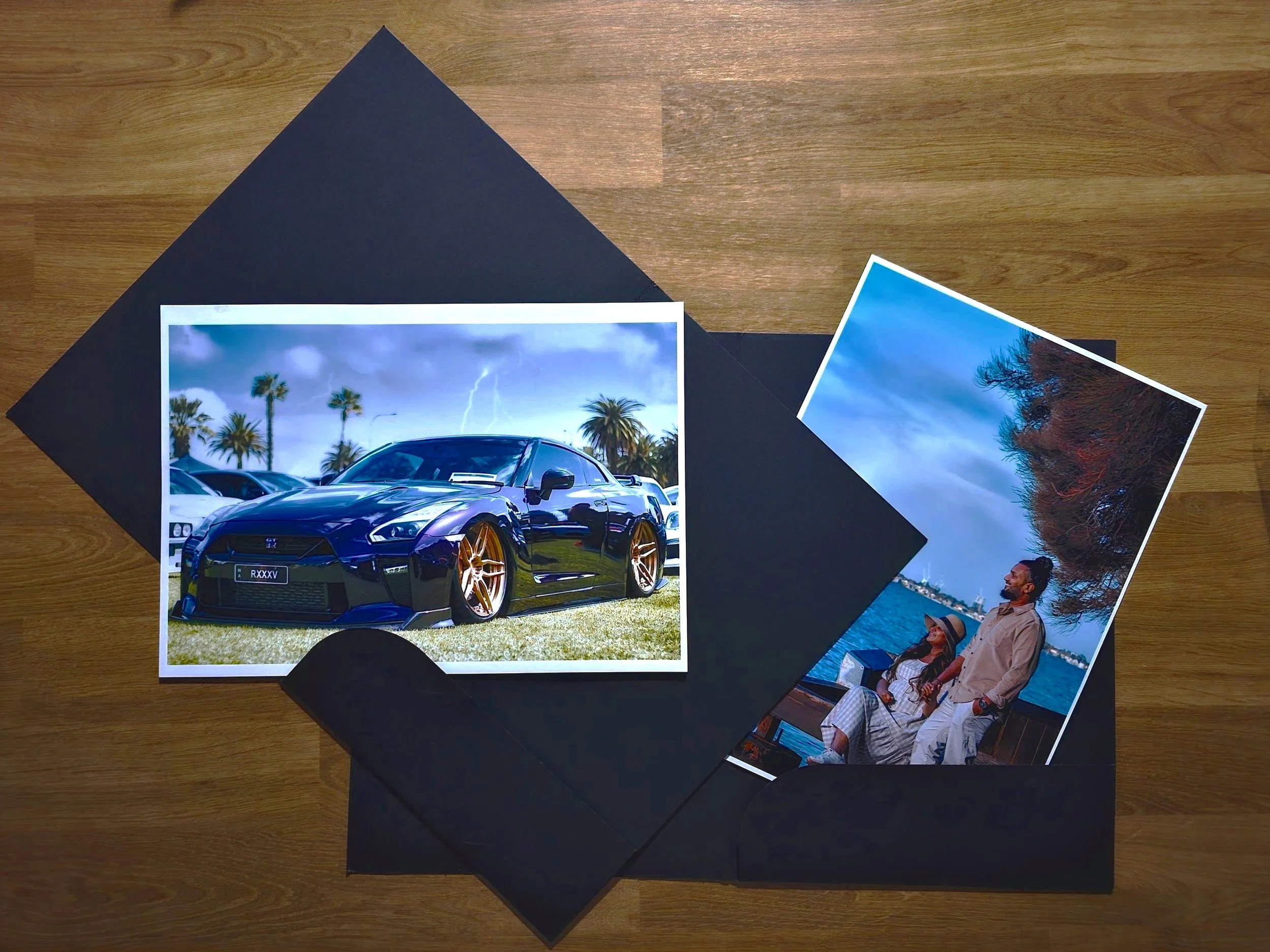 Two printed photographs in black envelopes on a wooden surface. One photo shows a black sports car with a lightning storm and palm trees in the background. The other photo depicts a man and woman enjoying a waterfront view with trees and water in the background.