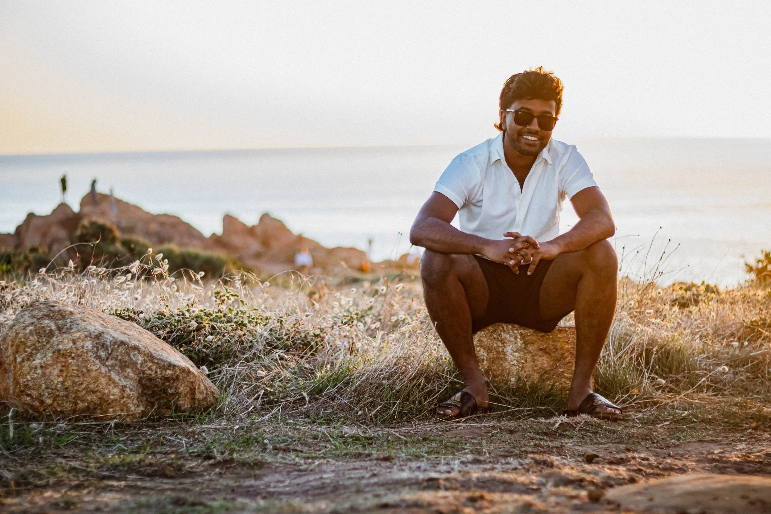 Young man wearing sunglasses, white shirt, and shorts squatting on a rock at the beach during sunset, smiling at the camera.