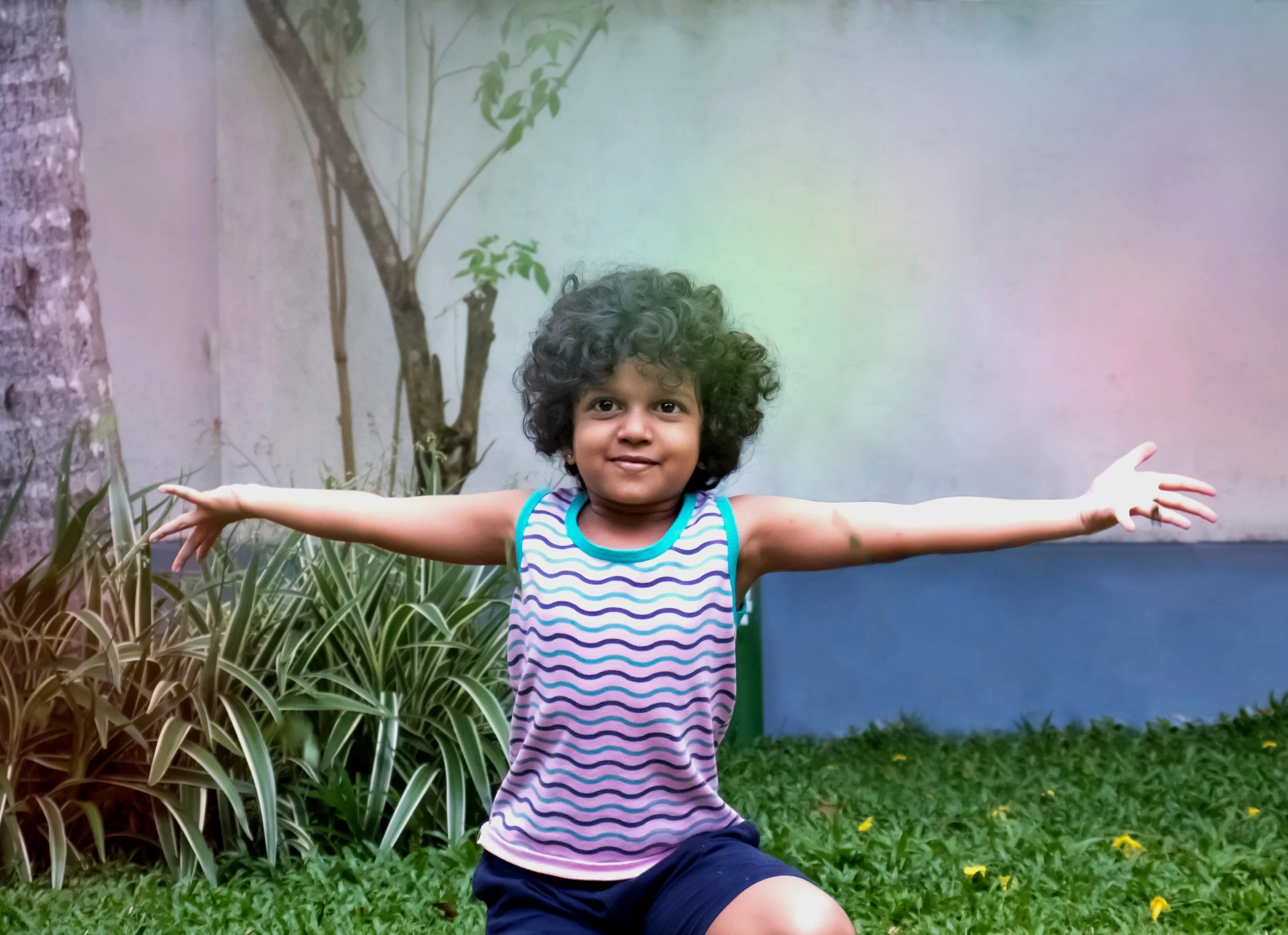 Young child with curly hair wearing a sleeveless striped shirt and shorts, standing outdoors with arms outstretched amidst green grass and plants.