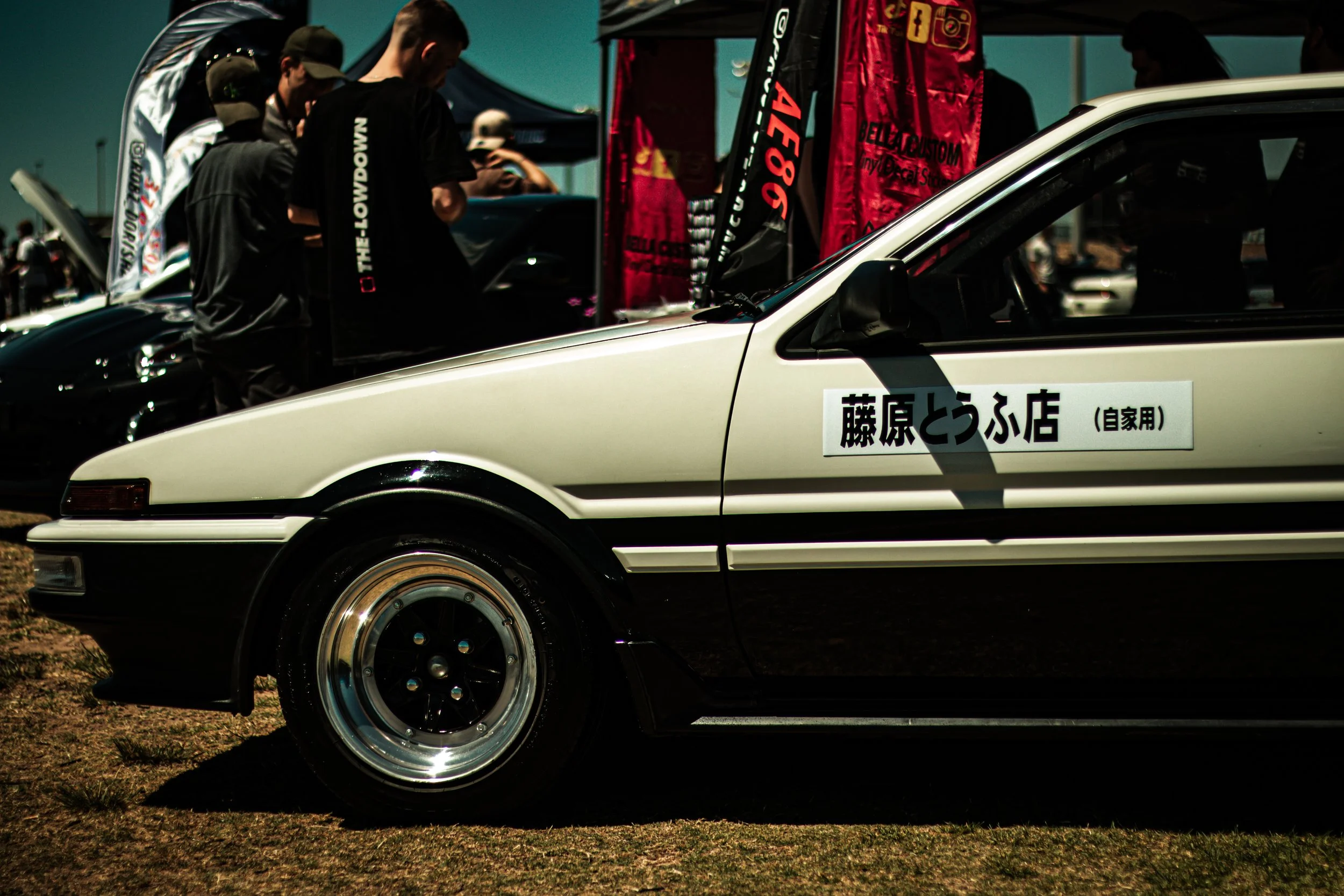 A white vintage car with Japanese writing on the side, parked at an outdoor car event with people and tents in the background.