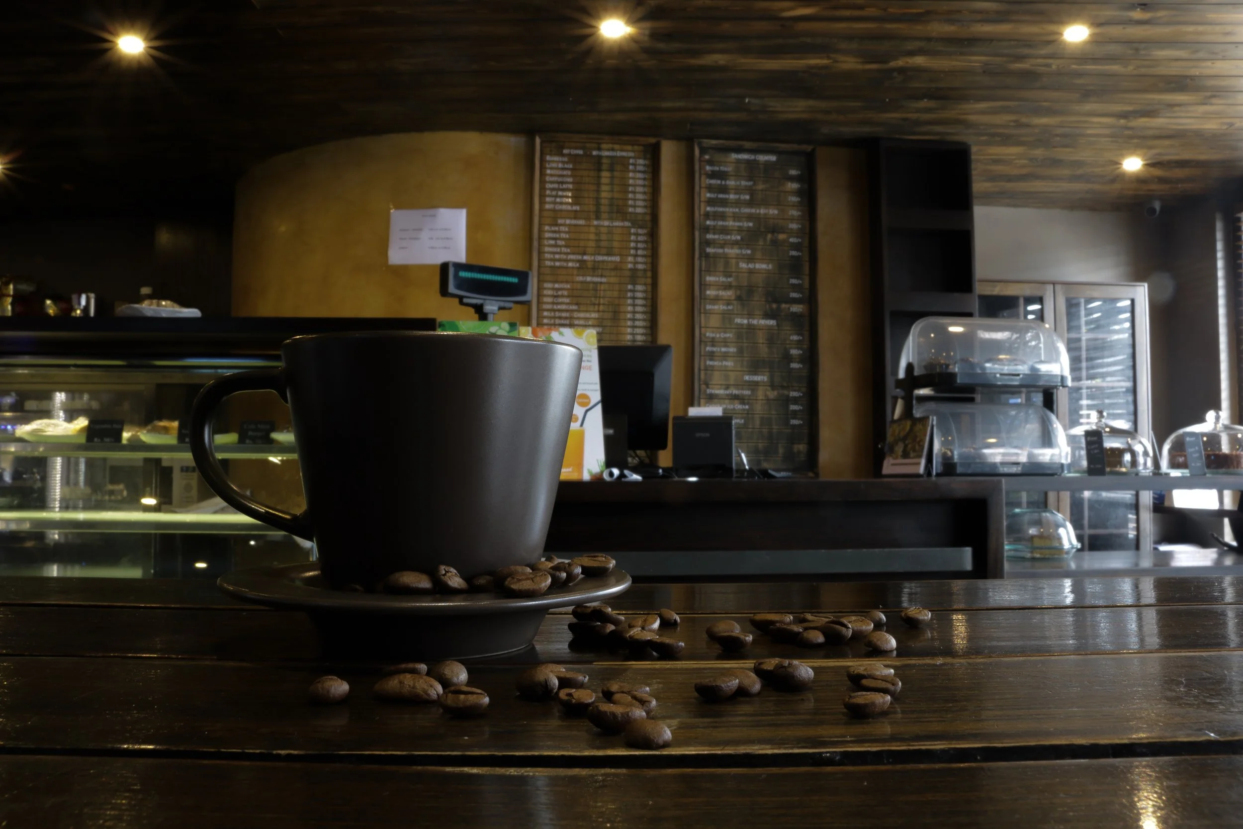 A black coffee cup on a saucer with scattered coffee beans on a wooden table inside a coffee shop, with a cafe counter, menu boards, and display cases in the background.