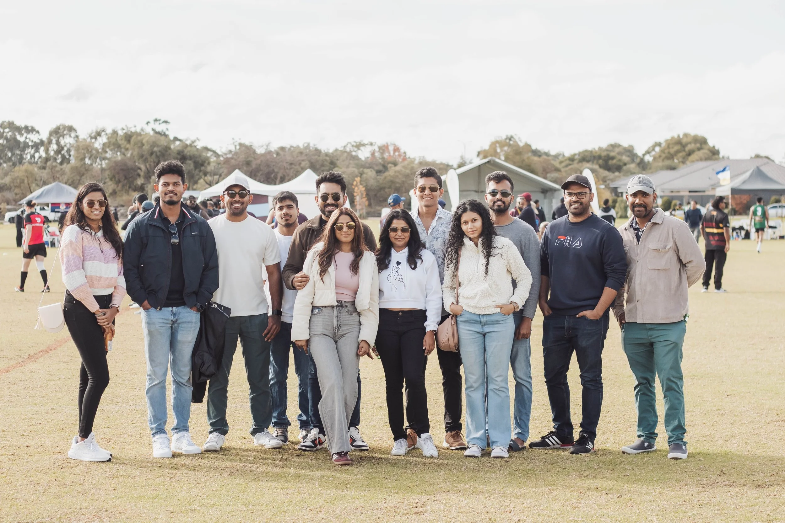 A group of fifteen people standing on a grassy field at an outdoor event, posing for a photo with tents, trees, and other people in the background.