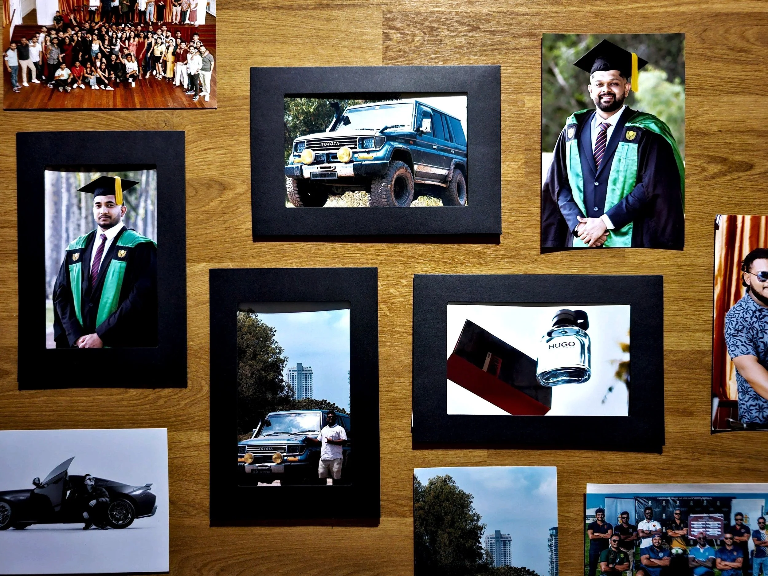 A collection of photographs on a wooden surface, including a group photo of people, a man in graduation attire, a blue off-road vehicle, a black sports car, a bottle of Hugo cologne, and other personal and scenic images.