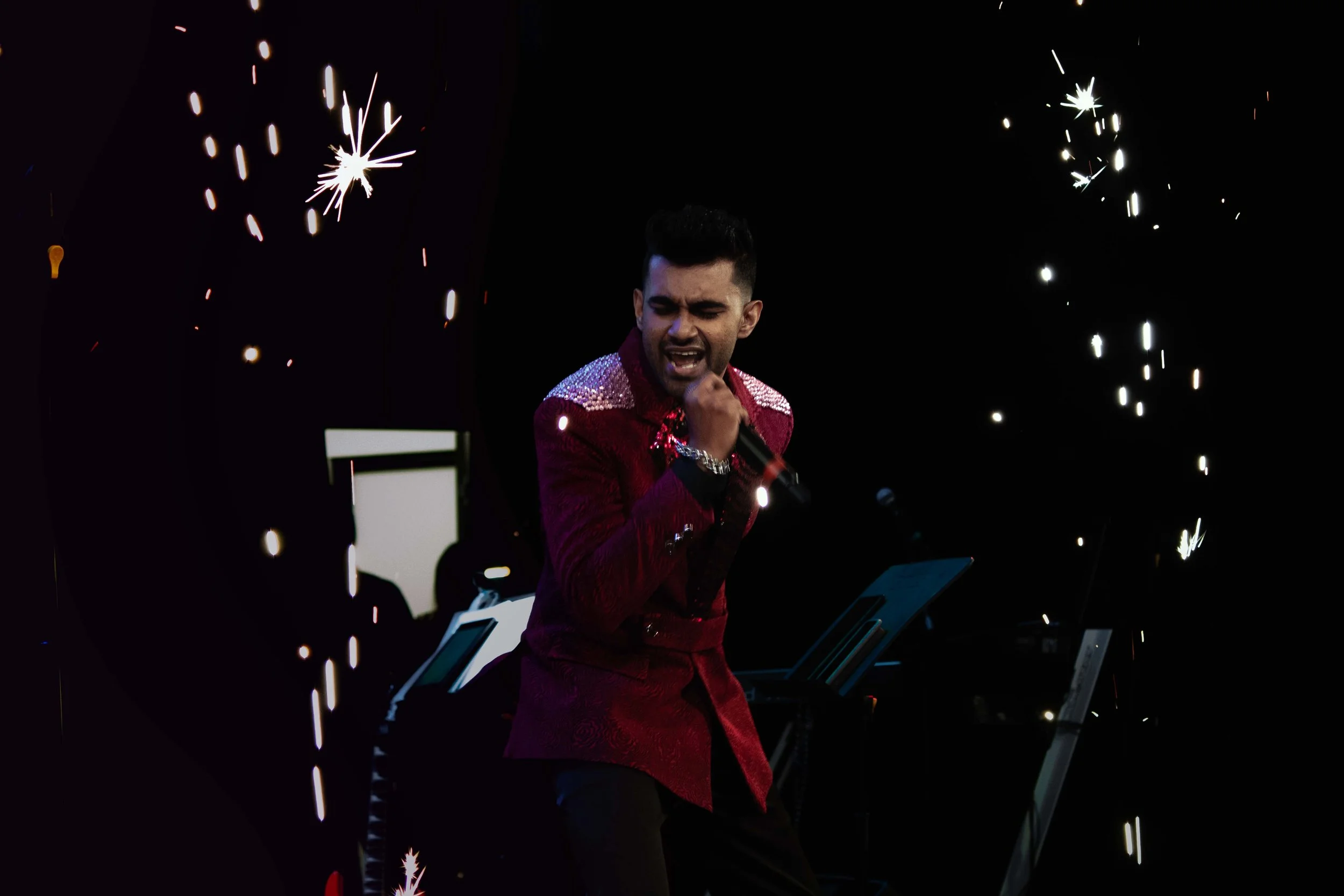 A man in a shiny red jacket sings passionately on stage, with sparklers creating bright streaks of light around him, and a keyboard visible in the background.