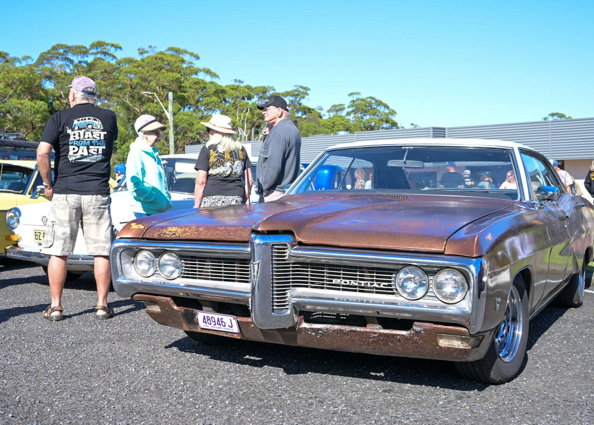 A group of older adults gathered around vintage cars at an outdoor car show on a sunny day.