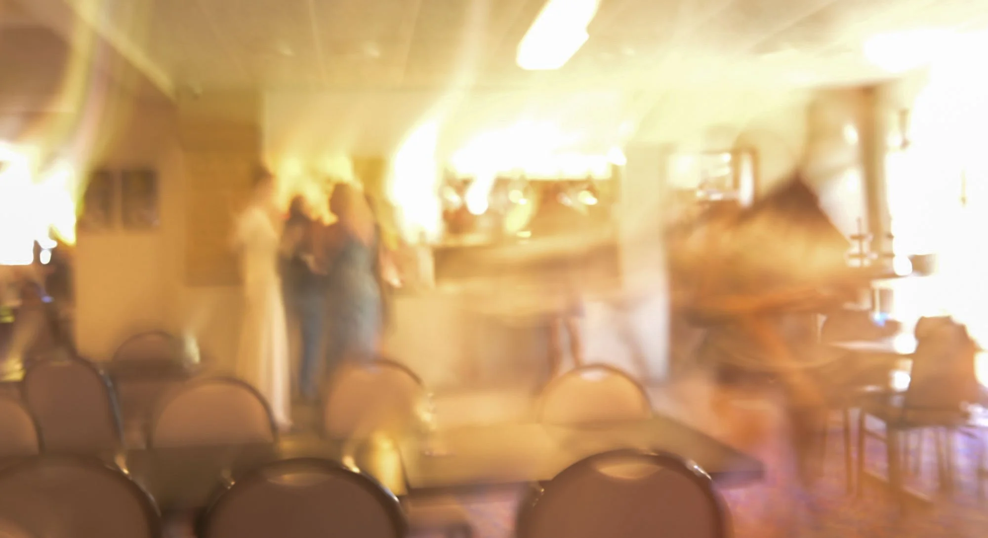 Dimly lit restaurant interior with some people standing near the bar area, wooden furniture, and multiple chairs in the foreground.