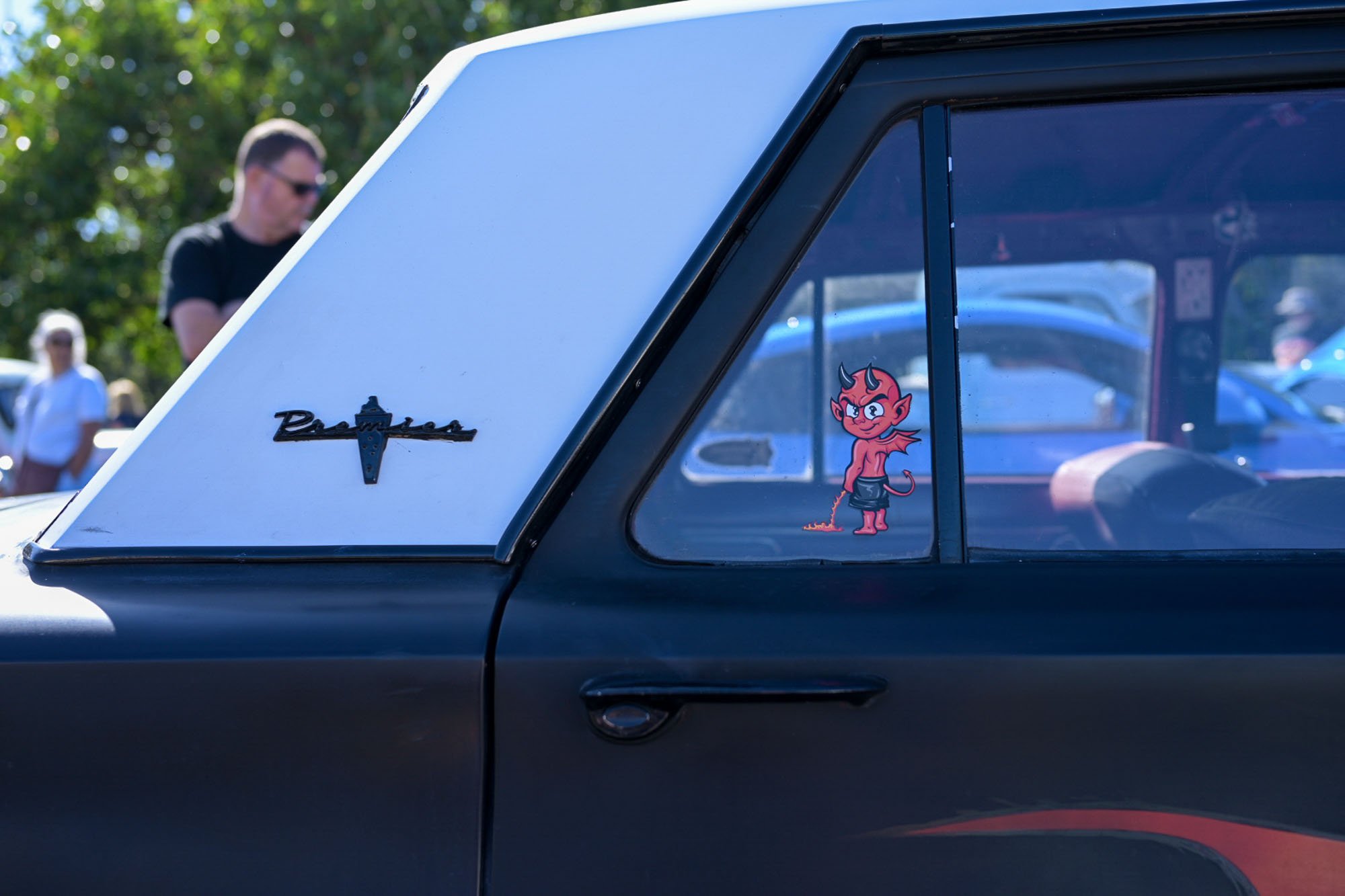 Close-up of a vehicle's side with a space-themed sticker of a cartoon devil character in sunglasses, with a painted badge reading 'Baldwin' and a vertical window showing reflections of the car's interior and outdoor scene.