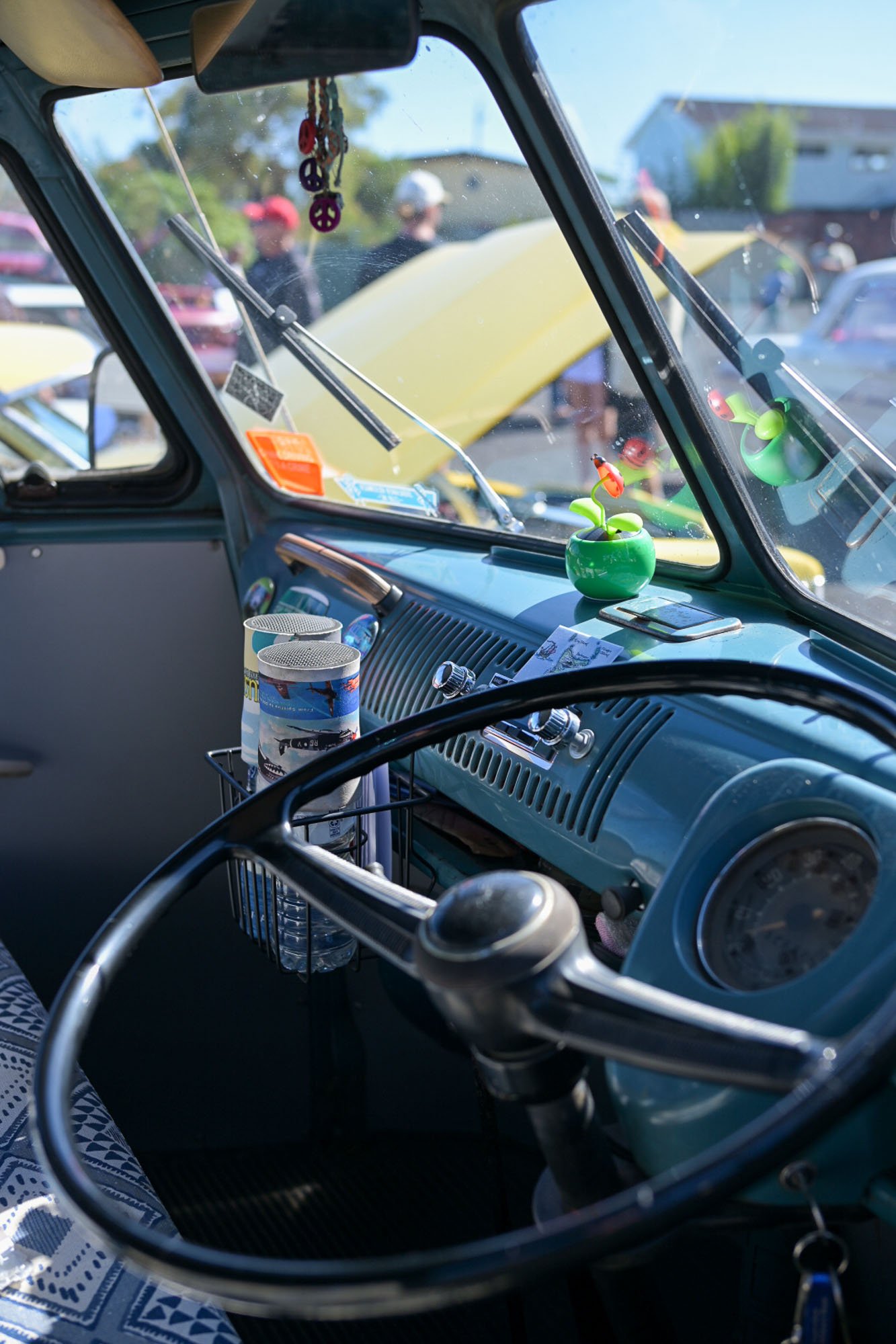 Inside view of a vintage car dashboard with a small green potted plant on the dashboard, several cans in a wire holder, and hanging jewelry charms, with reflections on the windshield showing people and other cars outside.