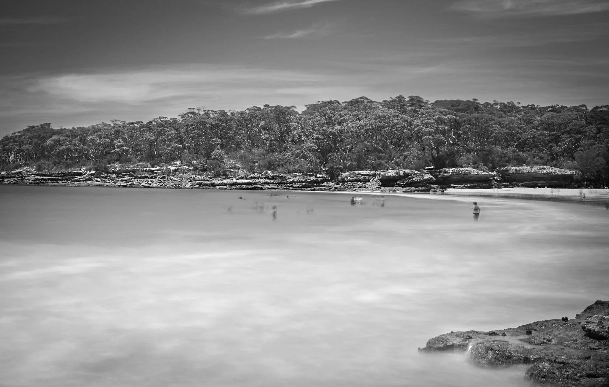 Black and white photo of a calm river with a rocky bank in the foreground, a dense tree-covered hillside in the background, and a sky with scattered clouds.