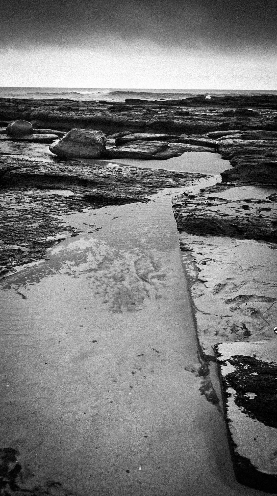 Black and white photo of a rocky shoreline with water and clouds in the sky.