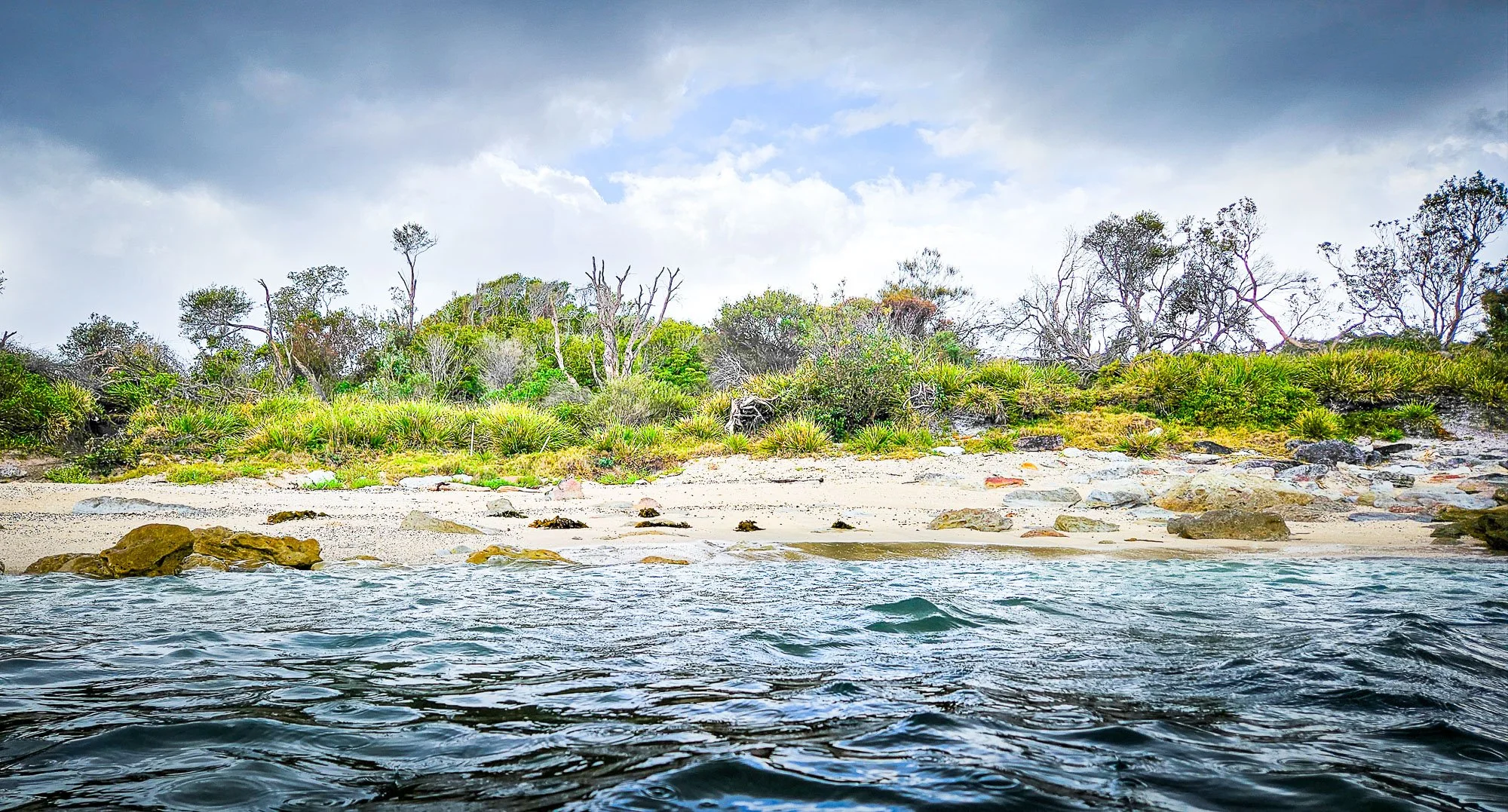 A coastal scene showing water in the foreground and a sandy beach with rocks, green bushes, and trees in the background under a cloudy sky.