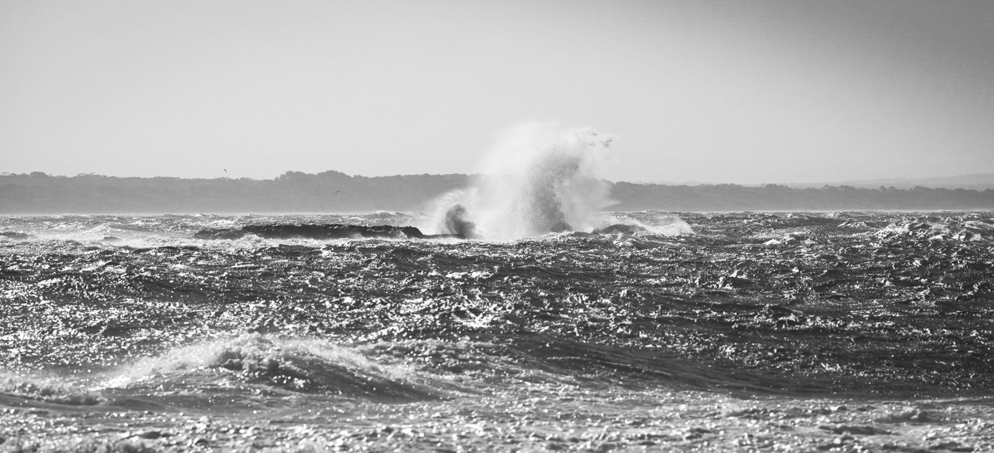 Black and white photo of ocean waves crashing against rocks with a distant shoreline and a cloudy sky.