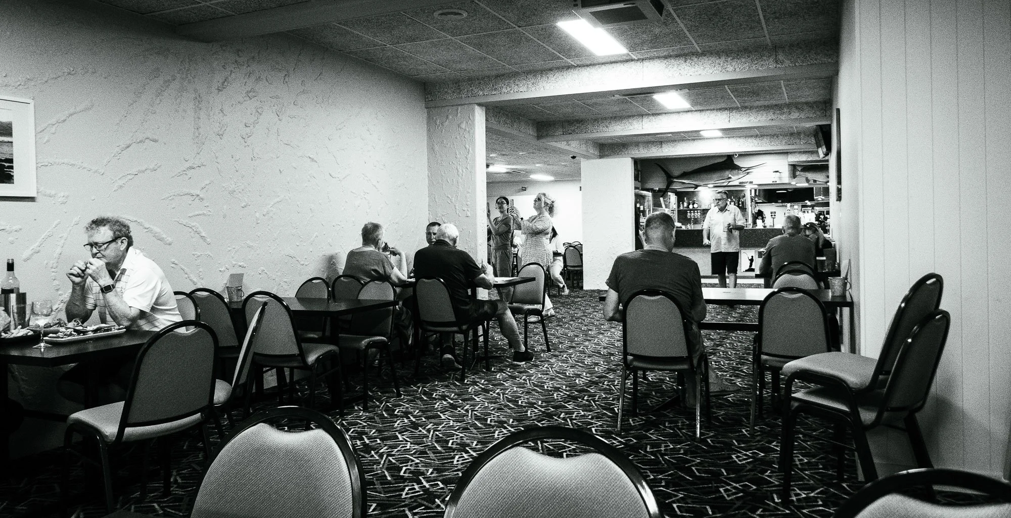 A black-and-white photo of a casual restaurant or bar with several patrons dining and socializing, some seated at tables and others standing near the bar in the background.