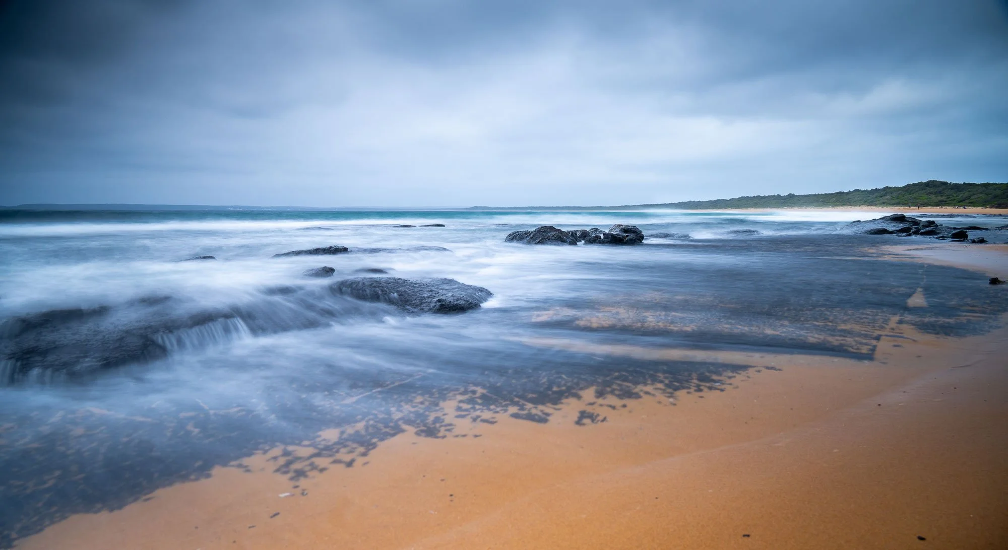 A long exposure photograph of a beach showing waves washing over rocks on the sandy shoreline with a cloudy sky overhead and a distant tree-lined coast.