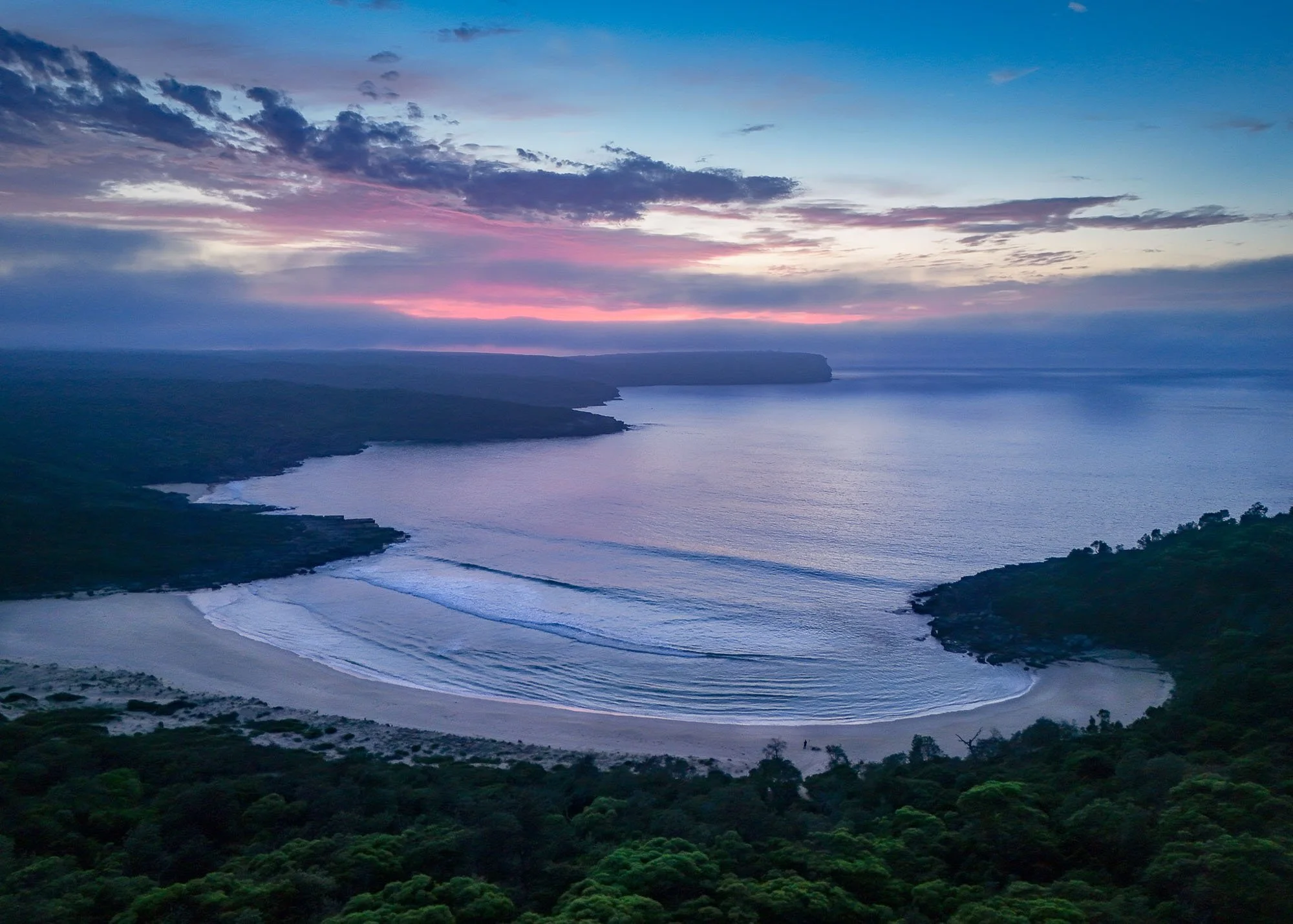 Sunset over a coastal landscape with dark clouds, rolling ocean waves, and hills covered in green vegetation.