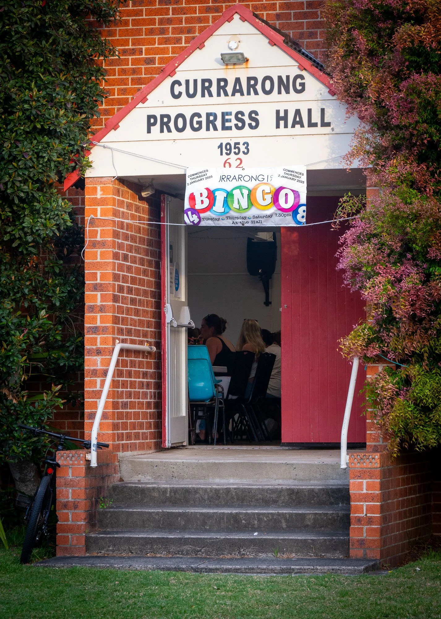 Red brick building entrance with a white triangular sign reading "Currarang Progress Hall 1953" and a crowd of people inside. A colorful banner with the word "BINGO" hangs above the entrance. Green bushes surround the doorway, and steps lead up to th