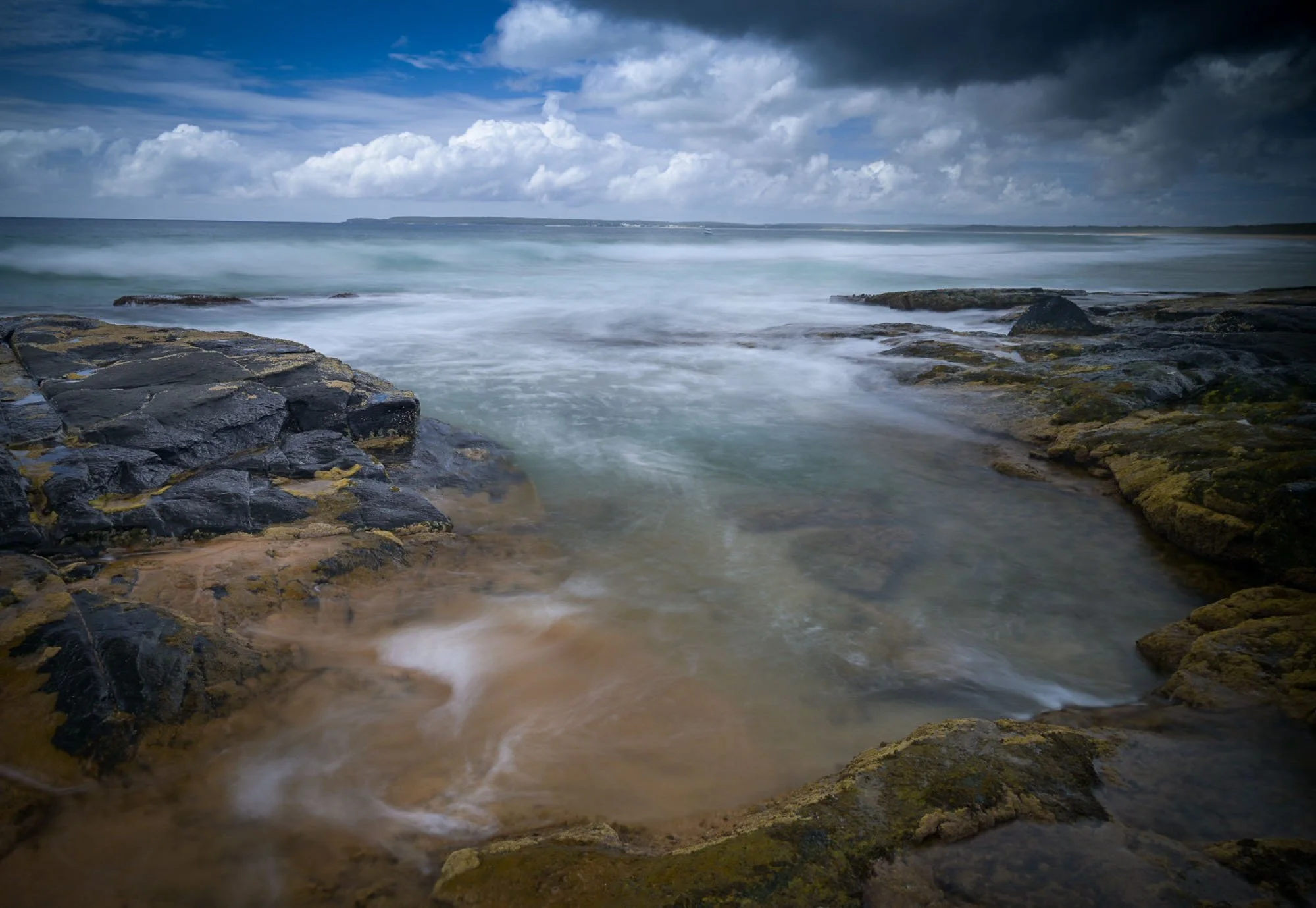 Coastal scene with rocky shoreline, waves crashing, dark clouds overhead, and distant land across the water.