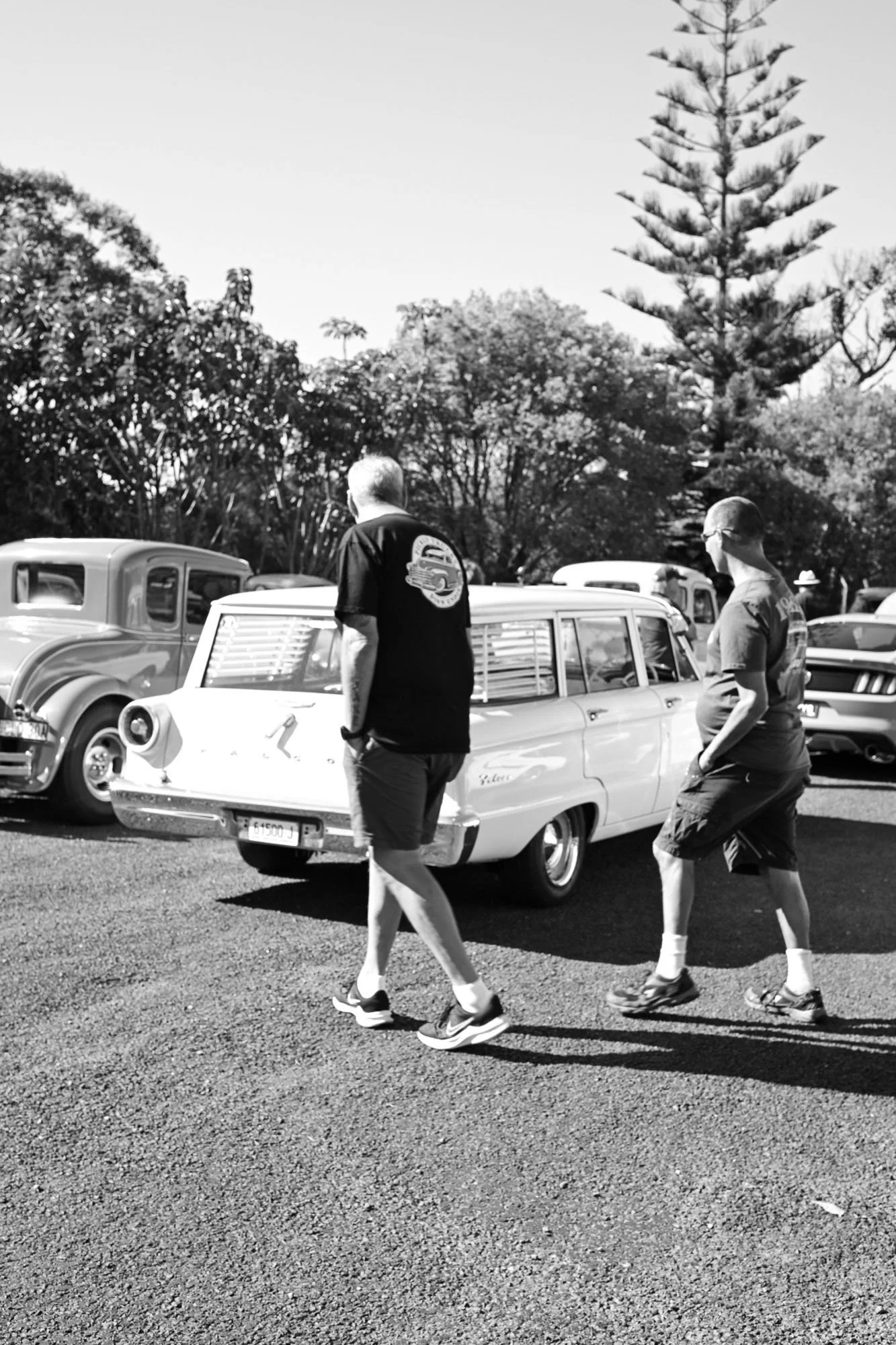 Black and white photo of people walking past classic cars at an outdoor event, with trees in the background.