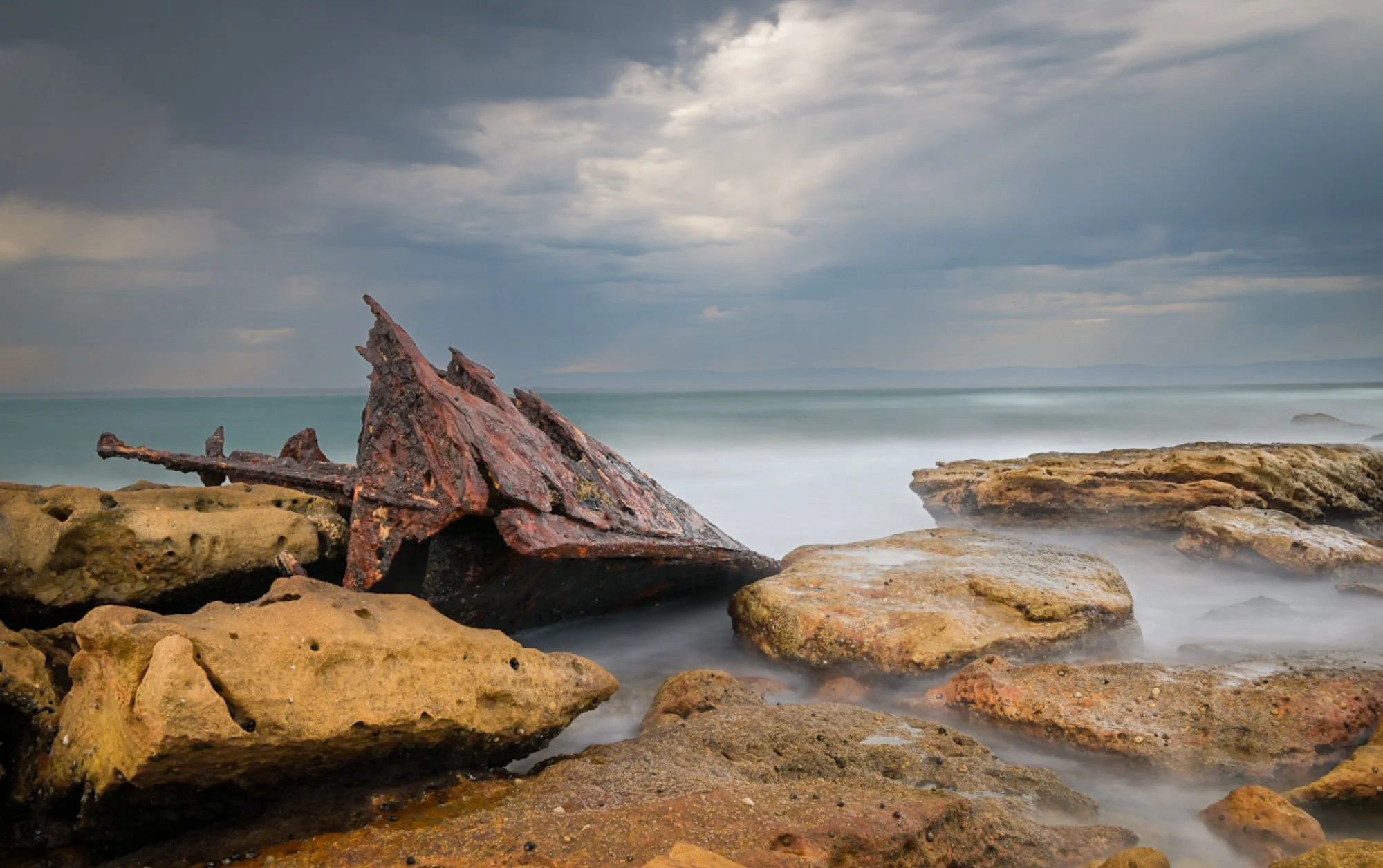 Rusted shipwreck on rocky shoreline under cloudy sky with ocean in the background.