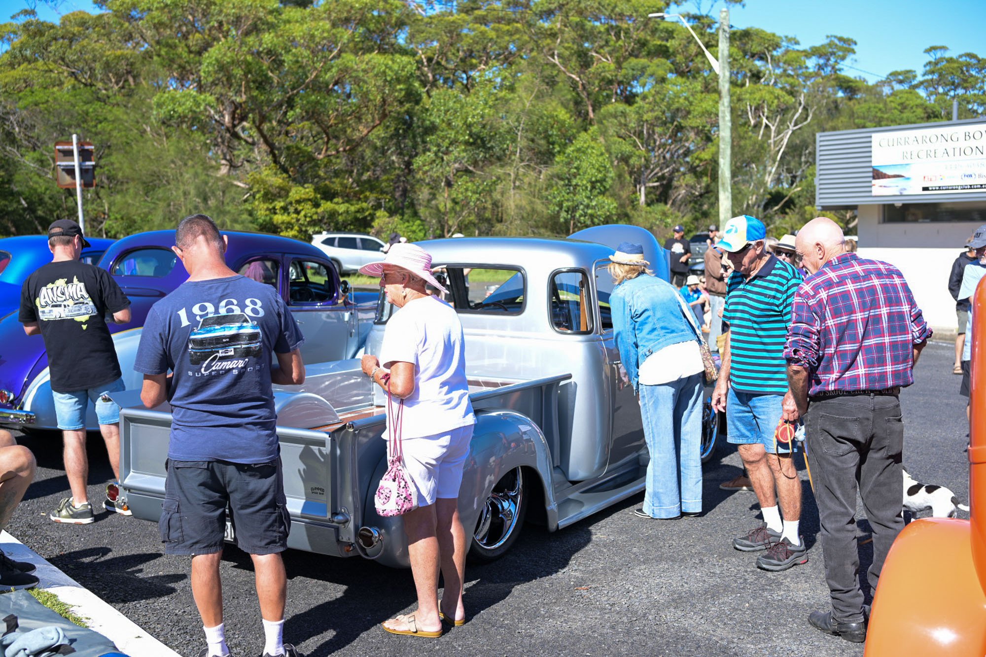 People gather around classic cars at an outdoor car show, with lush green trees and a billboard in the background.