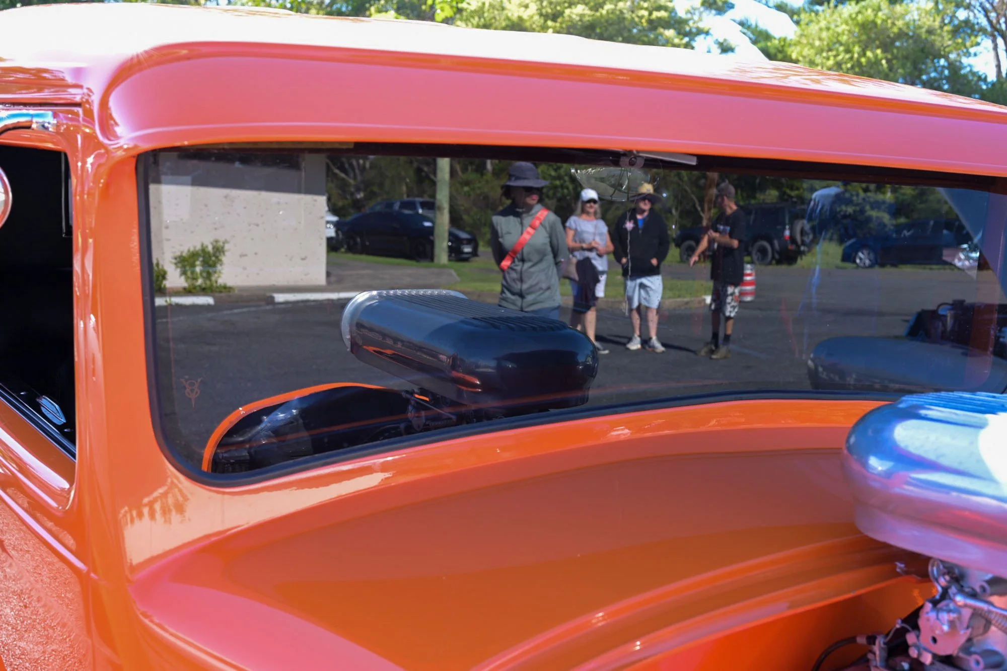 View of a group of people standing outside through the windshield of a vintage orange vehicle, with trees and parked cars in the background.