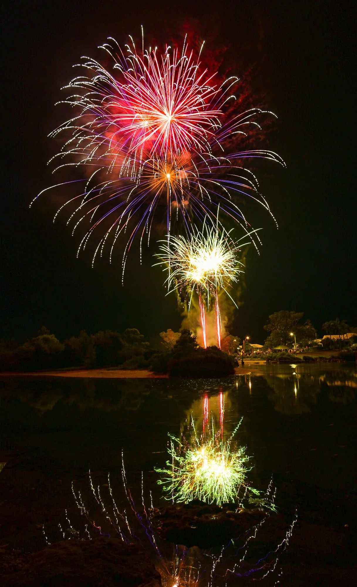Fireworks exploding in the night sky over a body of water with reflections of the fireworks.