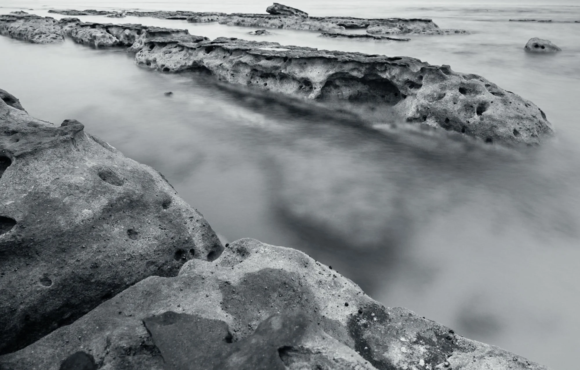 Black and white photo of rocks along a coastal shoreline with water flowing around them.