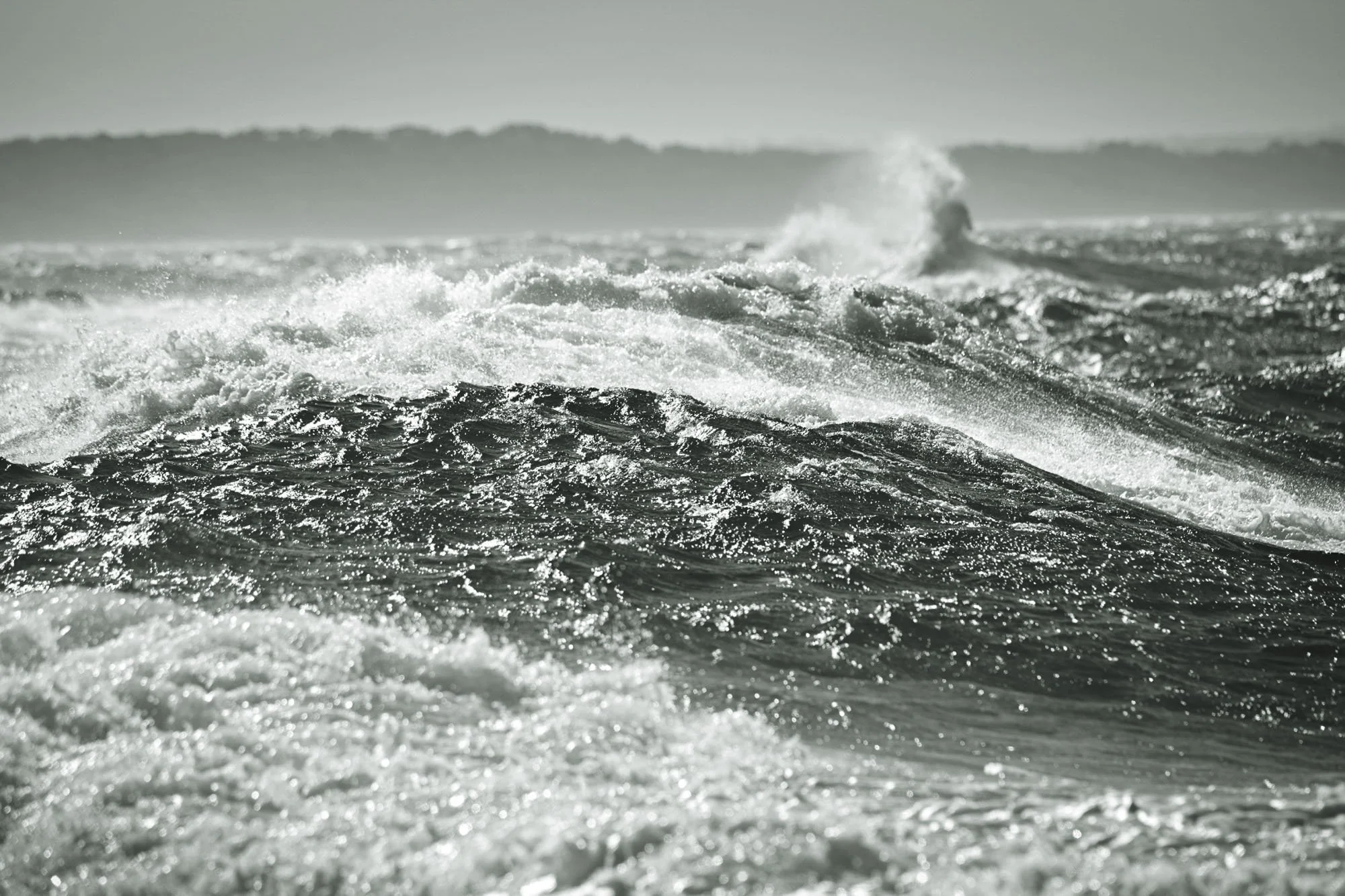 Black and white photo of ocean waves crashing with distant land or islands on the horizon.