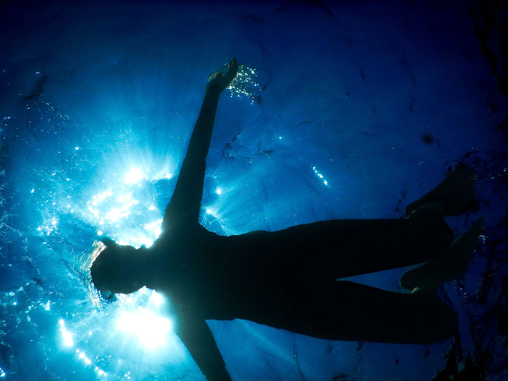 Person swimming underwater in a blue pool or ocean, with sunlight shining through the water and bubbles around.