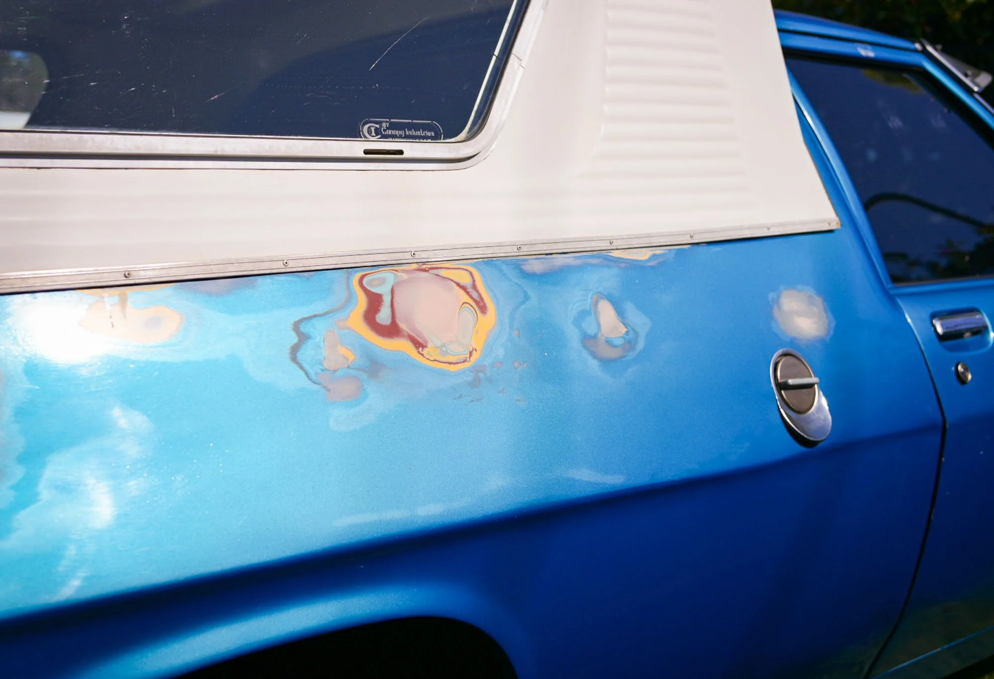 Close-up of a blue vintage car with a white roof showing a dent with multicolored paint damage.