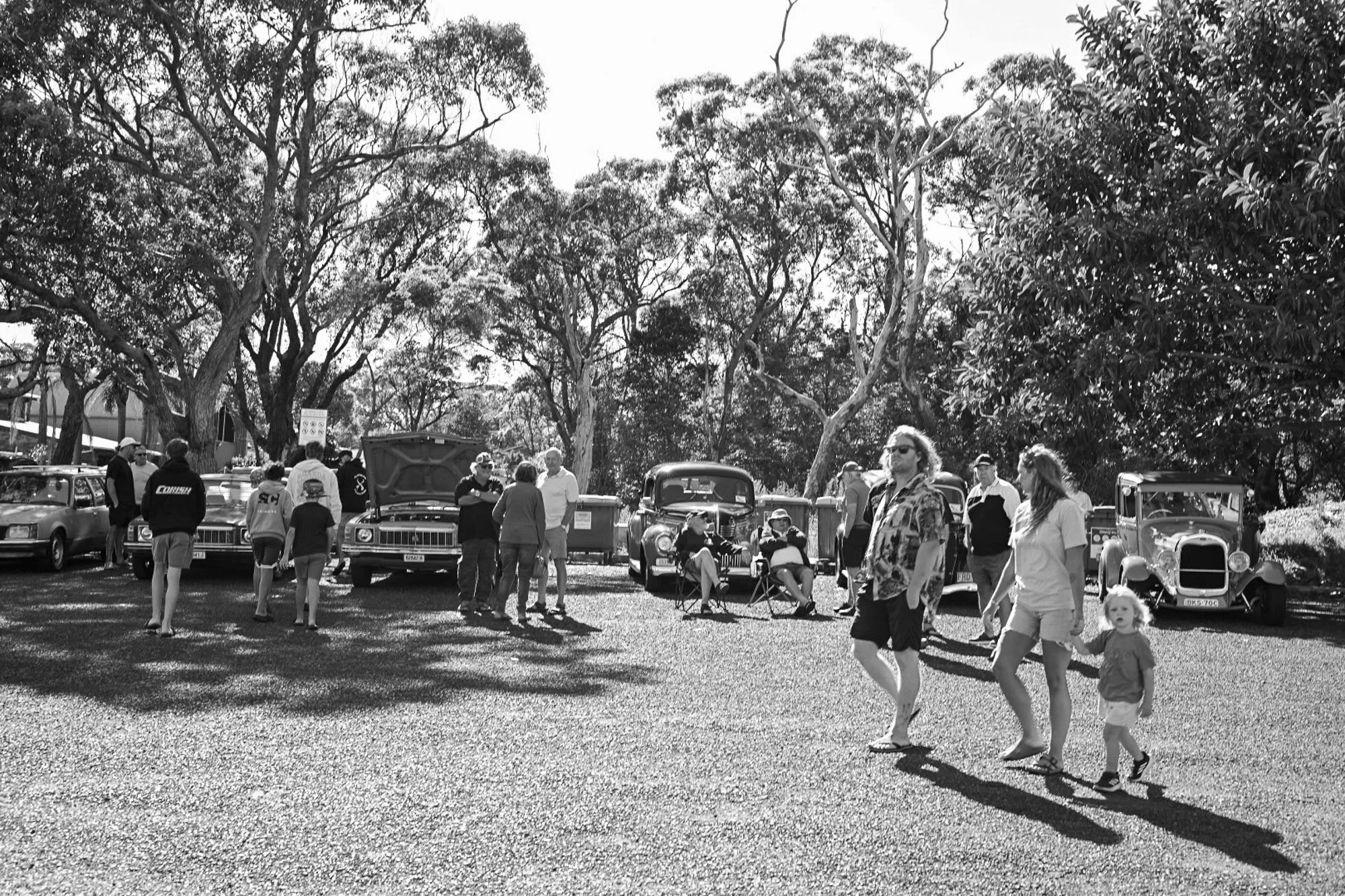 A black and white photo of people at an outdoor event with trees and vintage cars in the background.